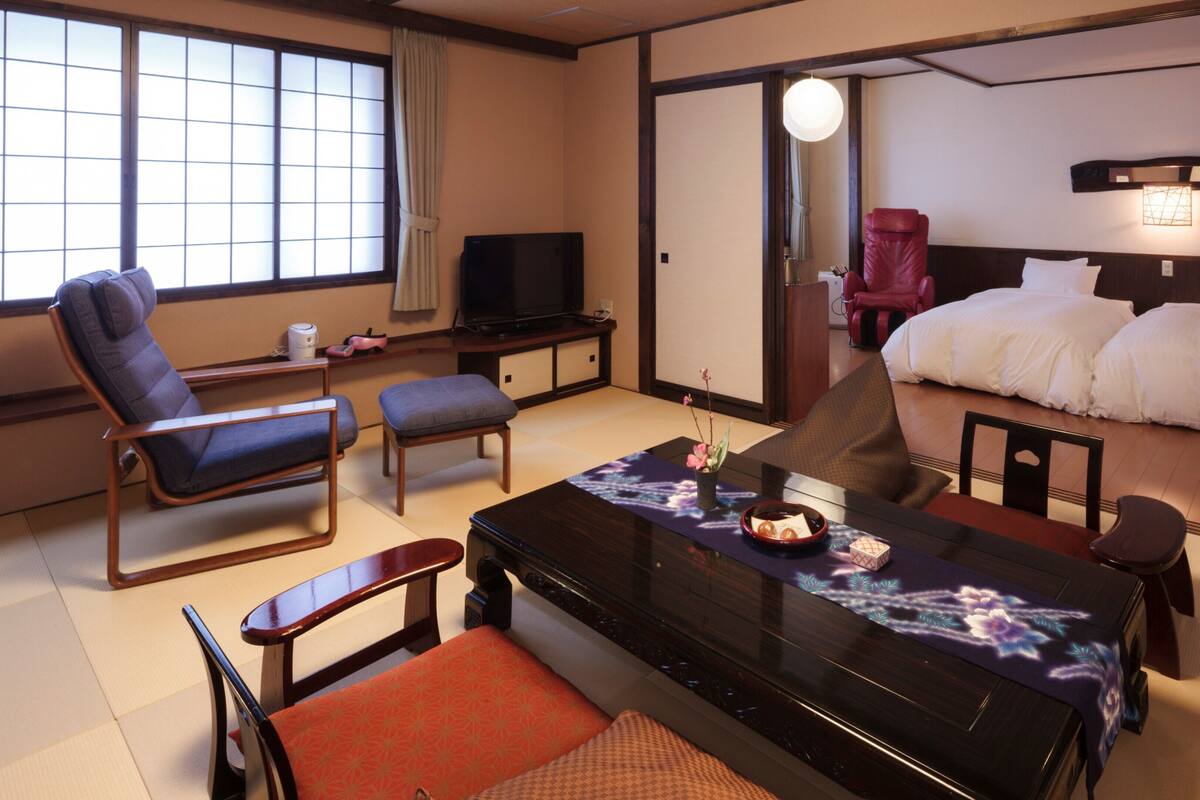 A hotel room with tatami flooring, a bed, a desk with a television, a chair, and a table with a decorative mat.