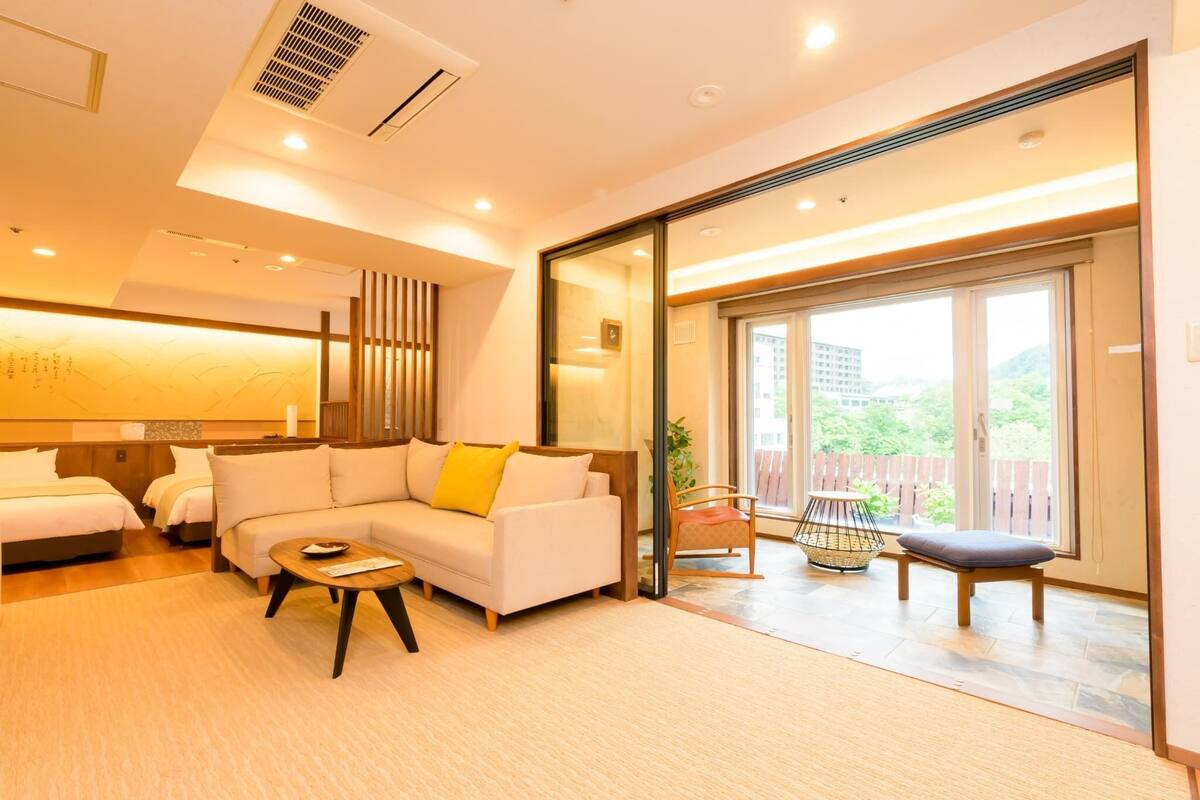 A modern hotel room with a sofa, a small table, a chair, and a large sliding glass door leading to an outdoor area.