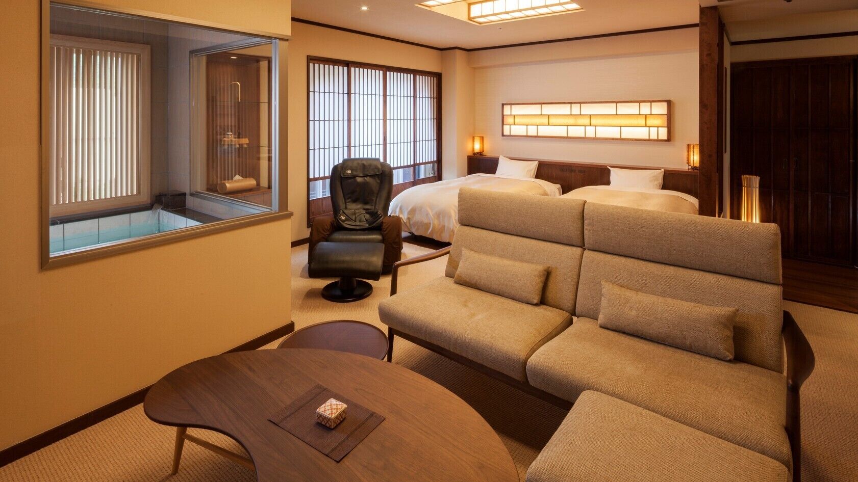 Une chambre d’hôtel équipée d’un canapé, d’un fauteuil, d’un lit, d’une table de chevet et d’une fenêtre avec des stores.