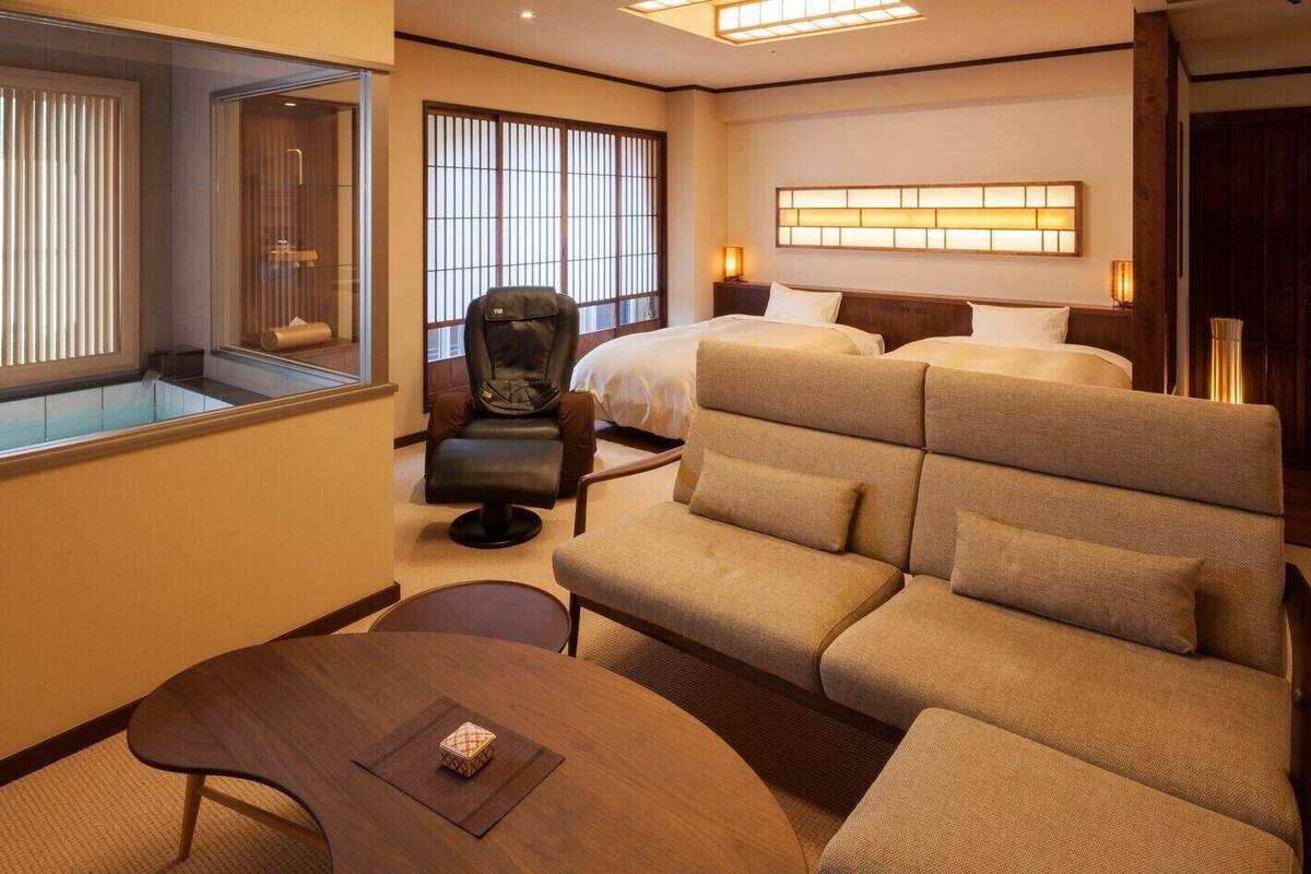 A hotel room with a sofa, a chair, a bed, a bedside table, and a window with blinds.