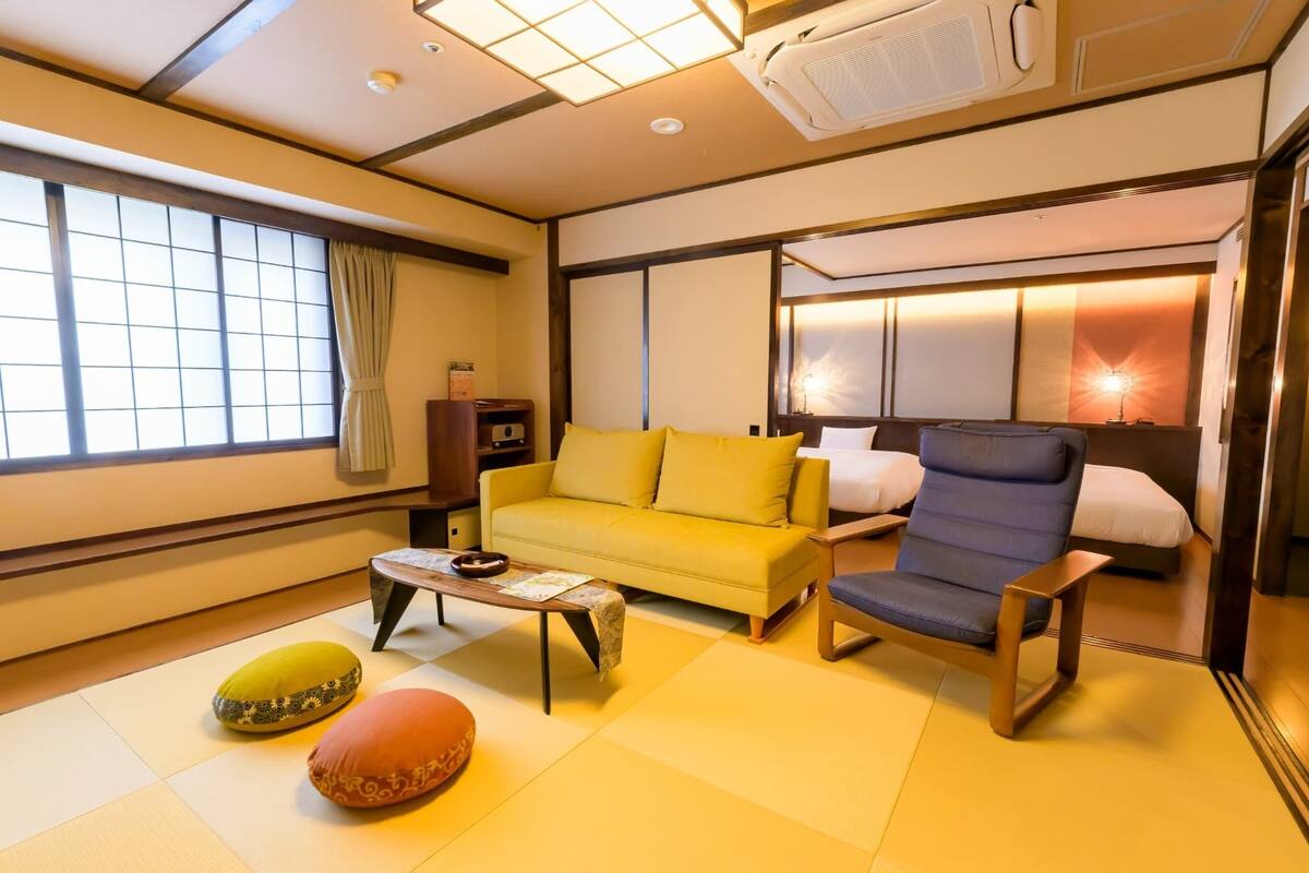 A hotel room with a sofa, armchair, and a bed with a headboard. There is a window with curtains, a small table with a tray, and a floor mat.