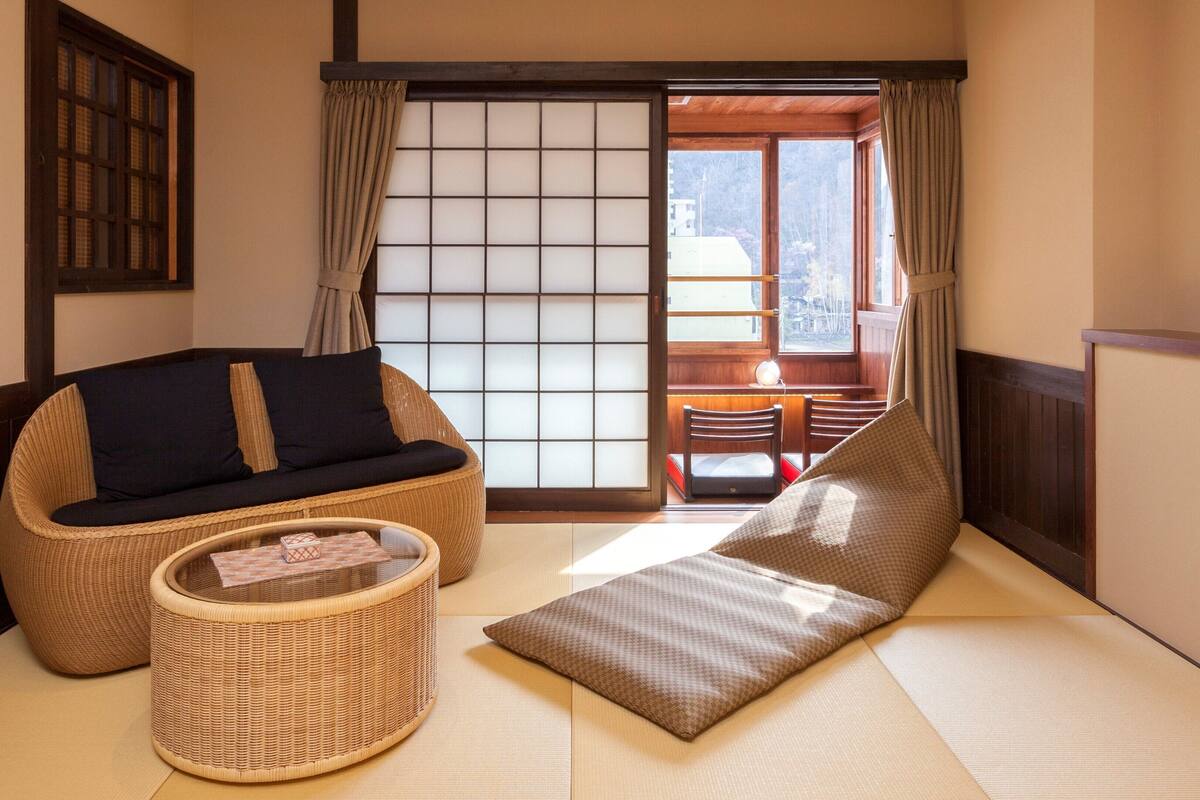 A traditional Japanese-style room with tatami flooring, a low table with a cushion, a woven chair, and a sliding door with a view of the outdoors.