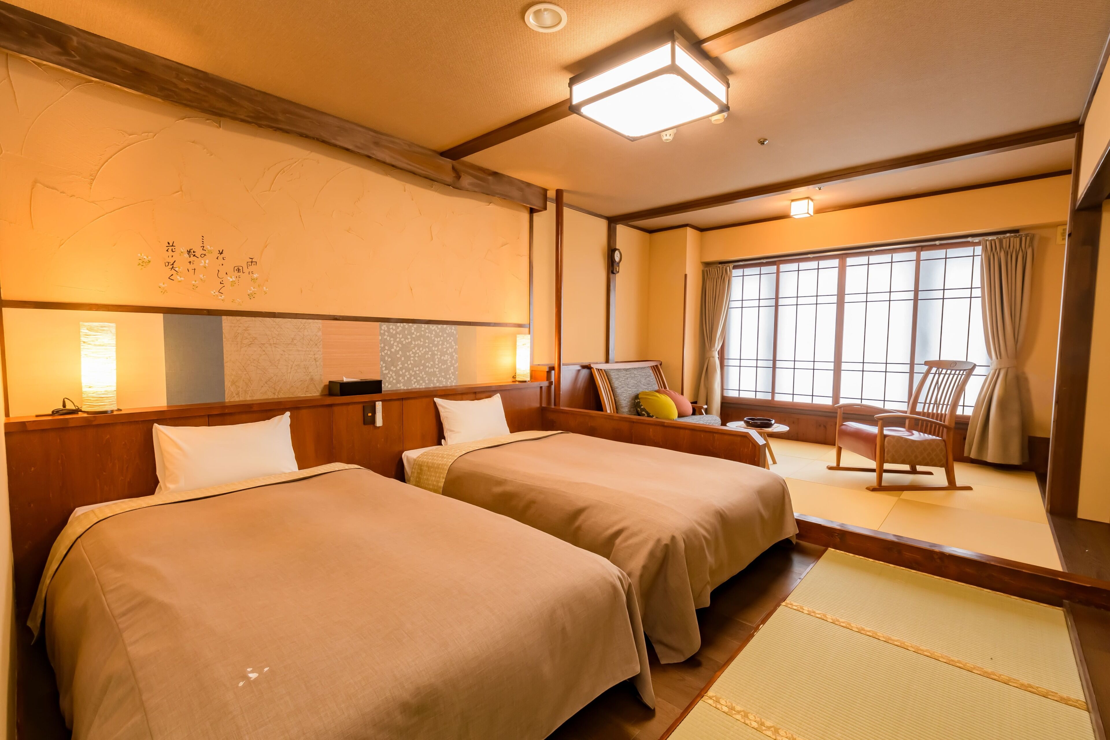 Une chambre d’hôtel avec deux lits, une tête de lit en bois, une grande fenêtre avec des rideaux, un sol traditionnel japonais recouvert de tatamis et une petite table avec une chaise.