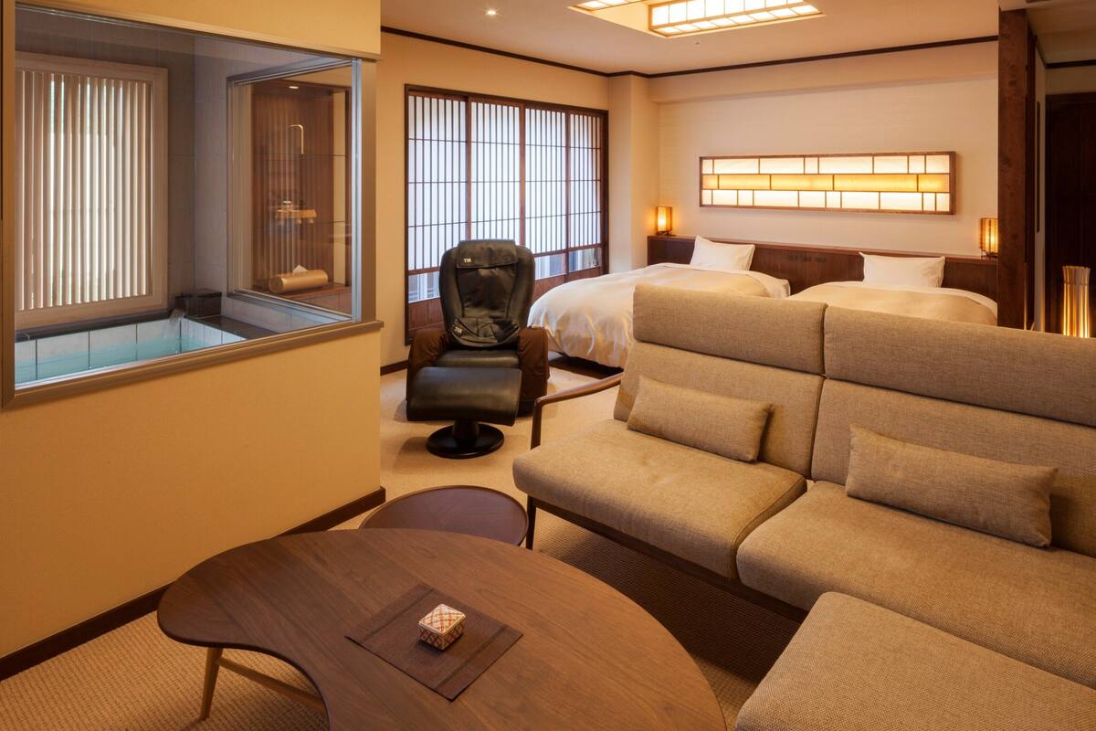 A hotel room with a sofa, a chair, a bed, a bedside table, and a window with blinds.
