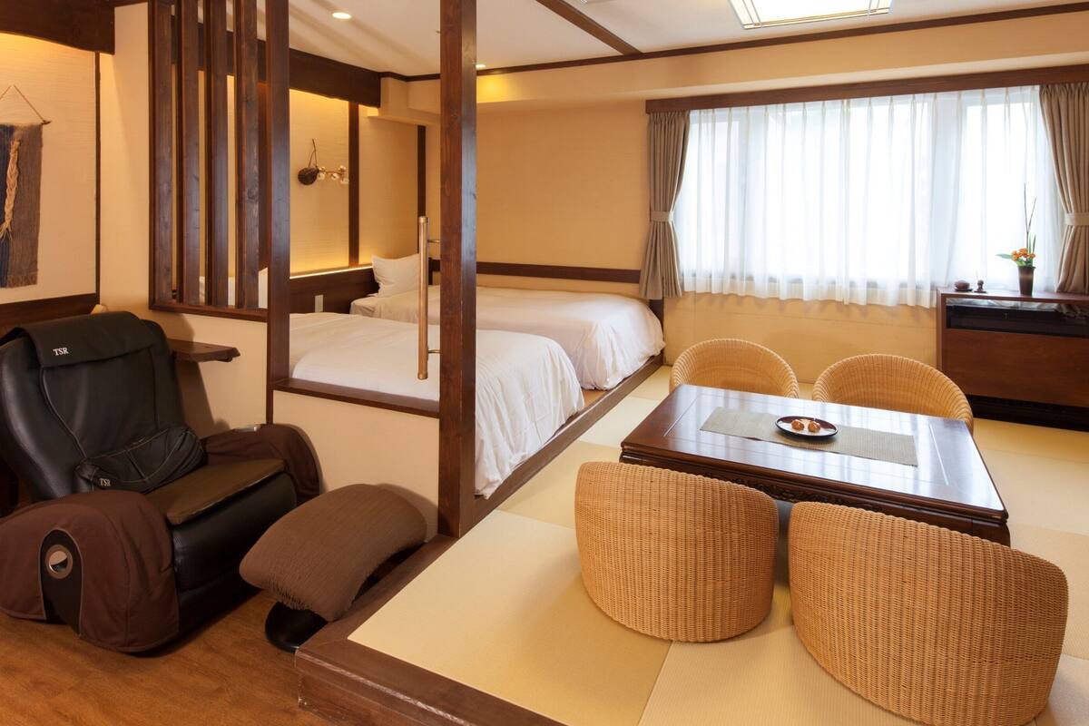 A hotel room with a bed, a sofa, a dining table with chairs, a massage chair, and a small table with a plate.