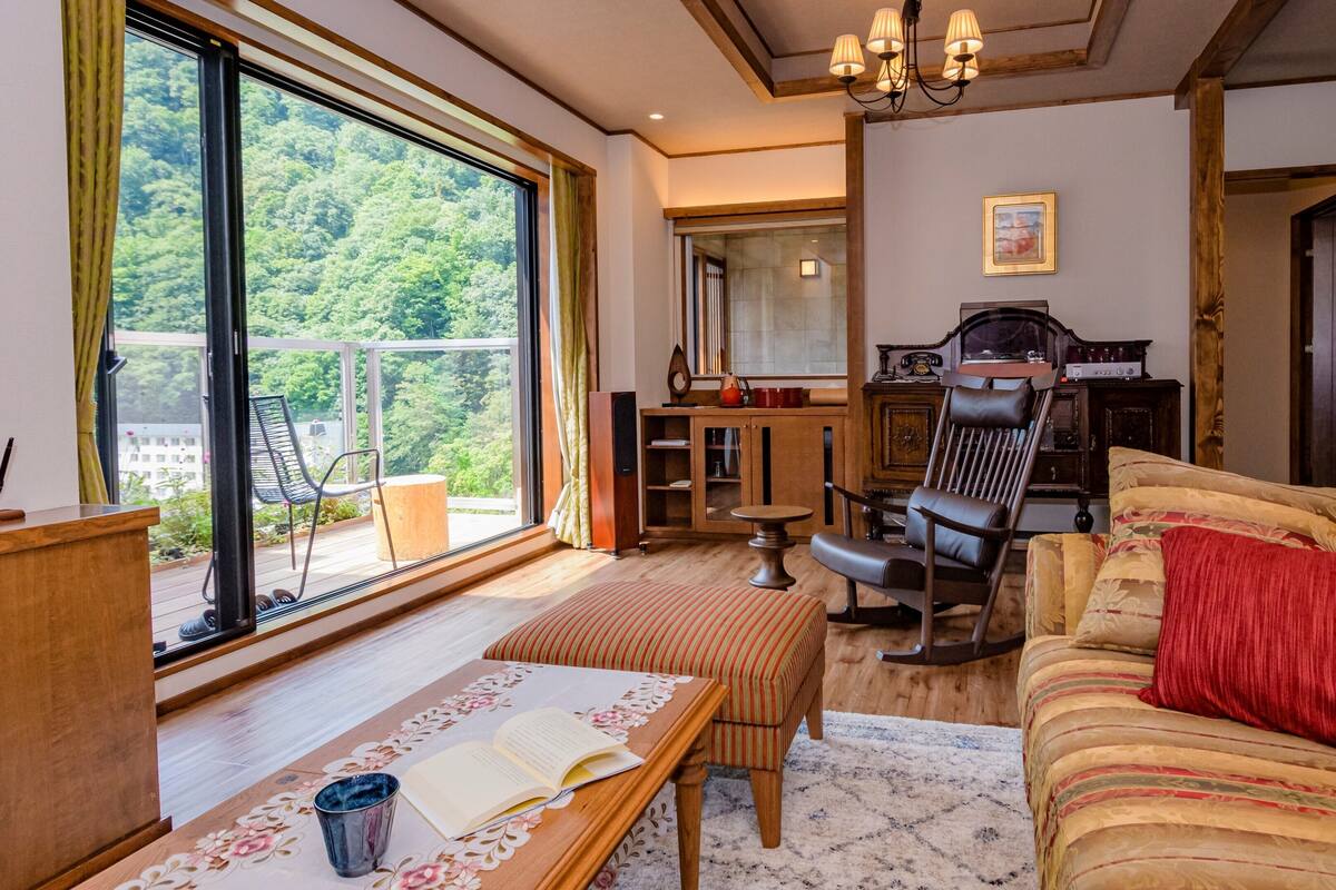 A living room with a sofa, a chair, a table with a book, a piano, and a view of greenery through large windows.
