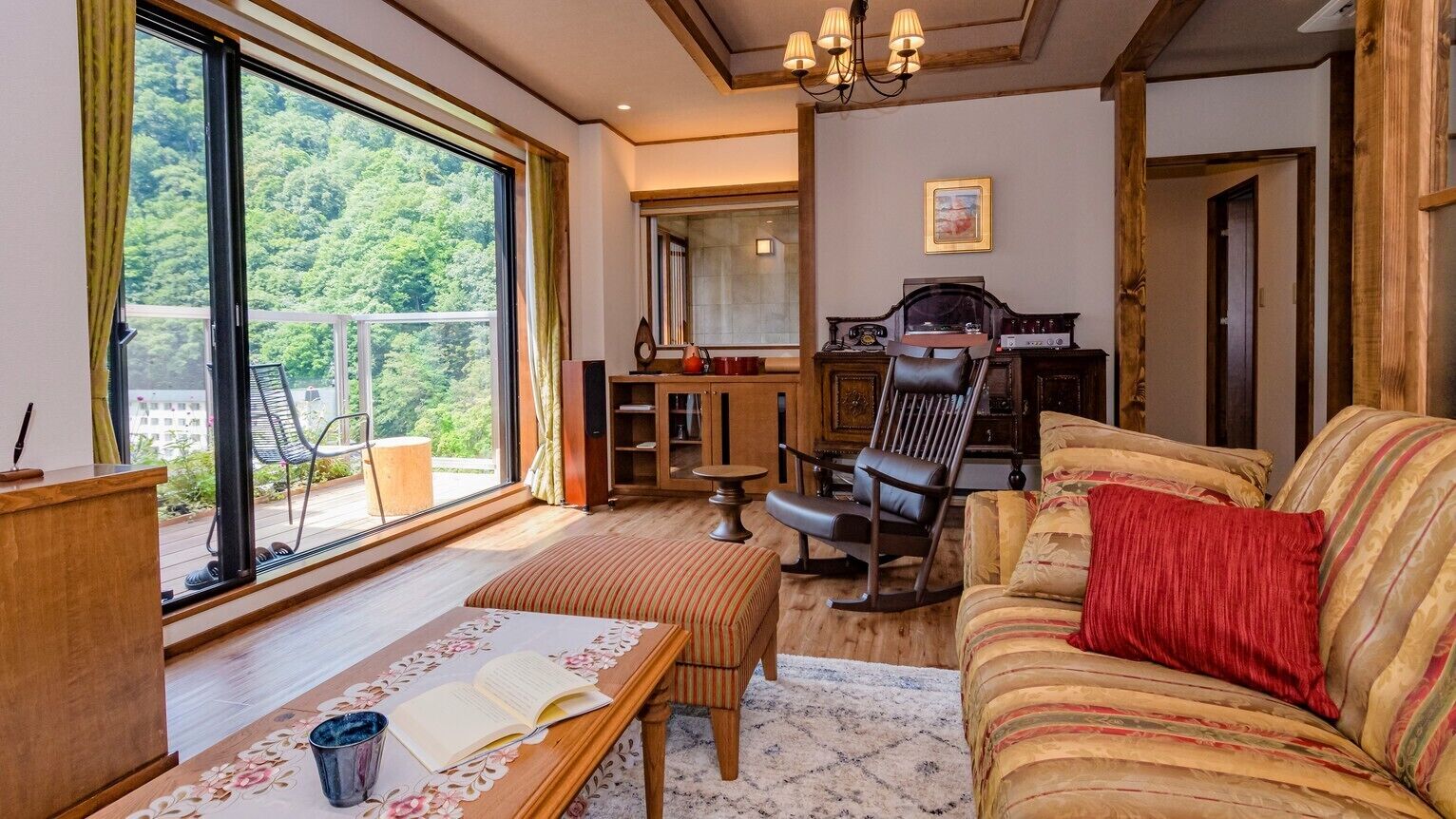 Un salon chaleureux avec une grande fenêtre, un canapé, un fauteuil à bascule et une table en bois sur laquelle repose un livre.