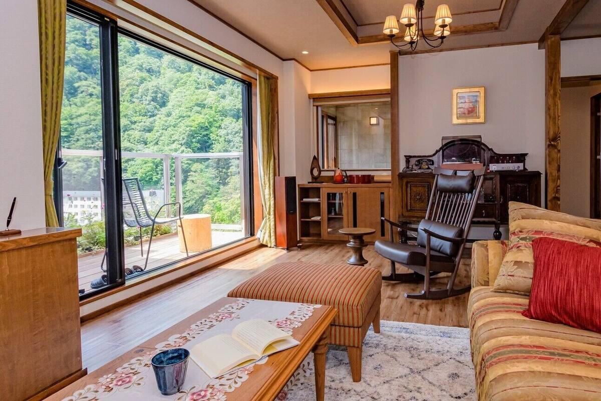 A cozy living room with a large window, a sofa, a rocking chair, and a wooden table with a book.