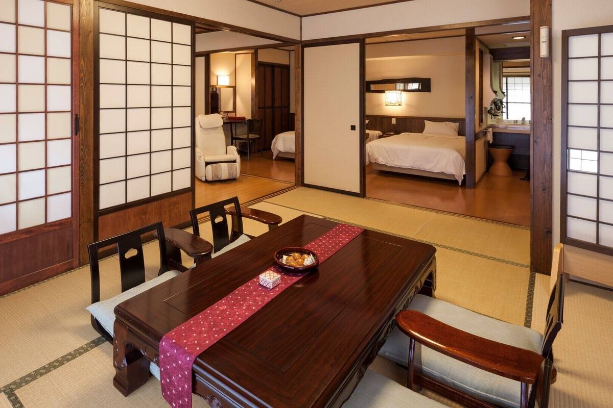 A traditional Japanese room with tatami flooring, a wooden dining table with a red placemat, and sliding doors leading to a bedroom.