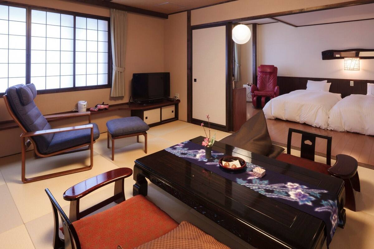 A hotel room with a bed, a desk with a television, a chair, a table with a decorative item, and tatami flooring.