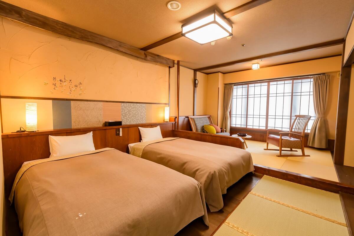 A traditional Japanese-style hotel room with two beds, tatami flooring, a wooden headboard, a large window with curtains, and a wooden rocking chair.