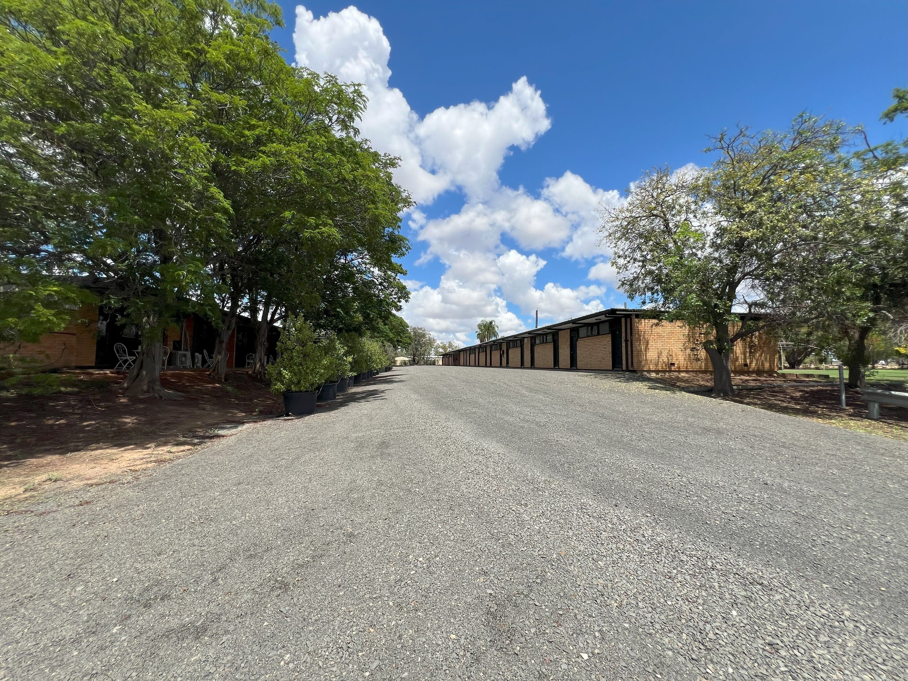 A gravel road leading to a row of brick buildings with large windows, surrounded by trees.