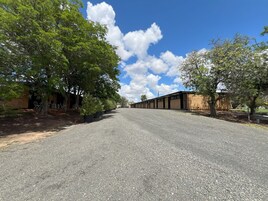A gravel road leading to a row of brick buildings with large windows, surrounded by trees.