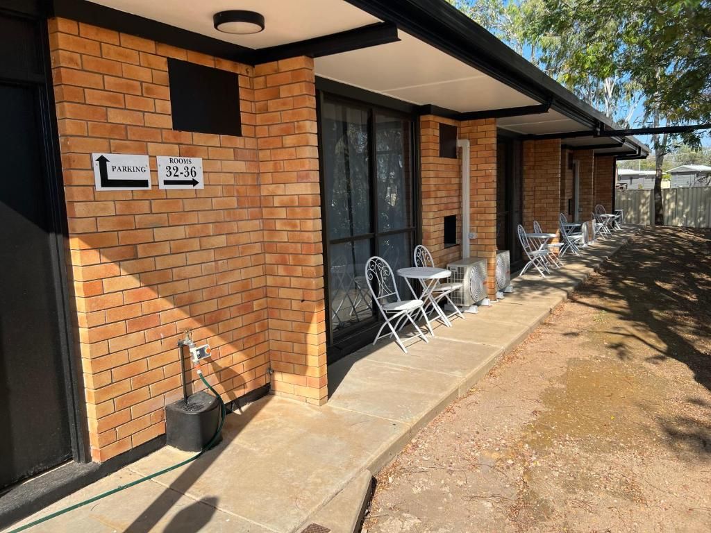 Un edificio con fachada de ladrillo, un área de estar al aire libre techada con mesas y sillas, y señalización que indica estacionamiento y números de habitaciones.