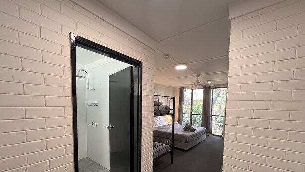 A corridor leading to a bedroom with a bunk bed, a bathroom with a glass shower enclosure, and a view of the outdoors through large windows. - Jumbuck Motel (Longreach)