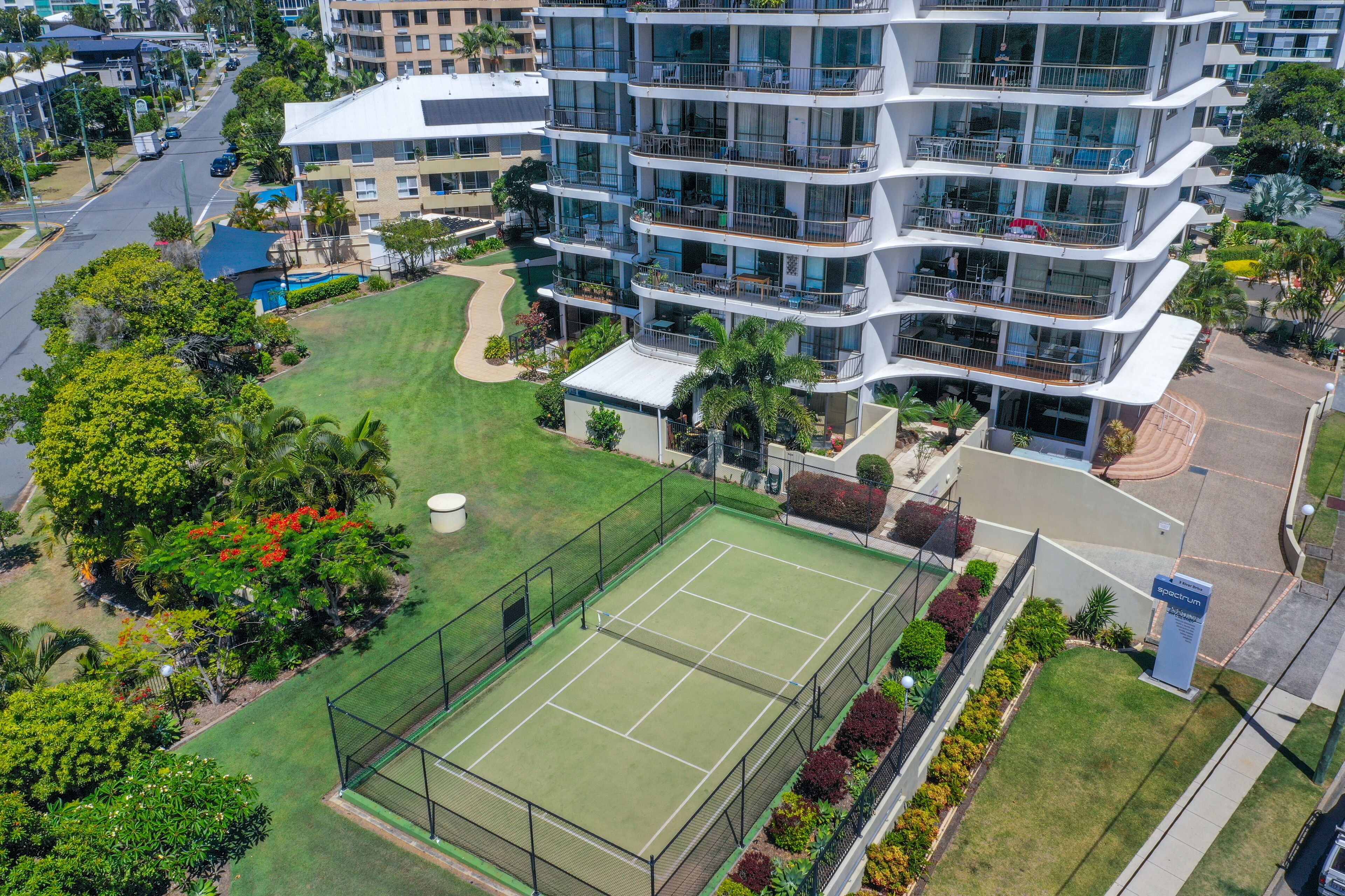 Un hotel de varios pisos con balcones, cancha de tenis y vegetación circundante.