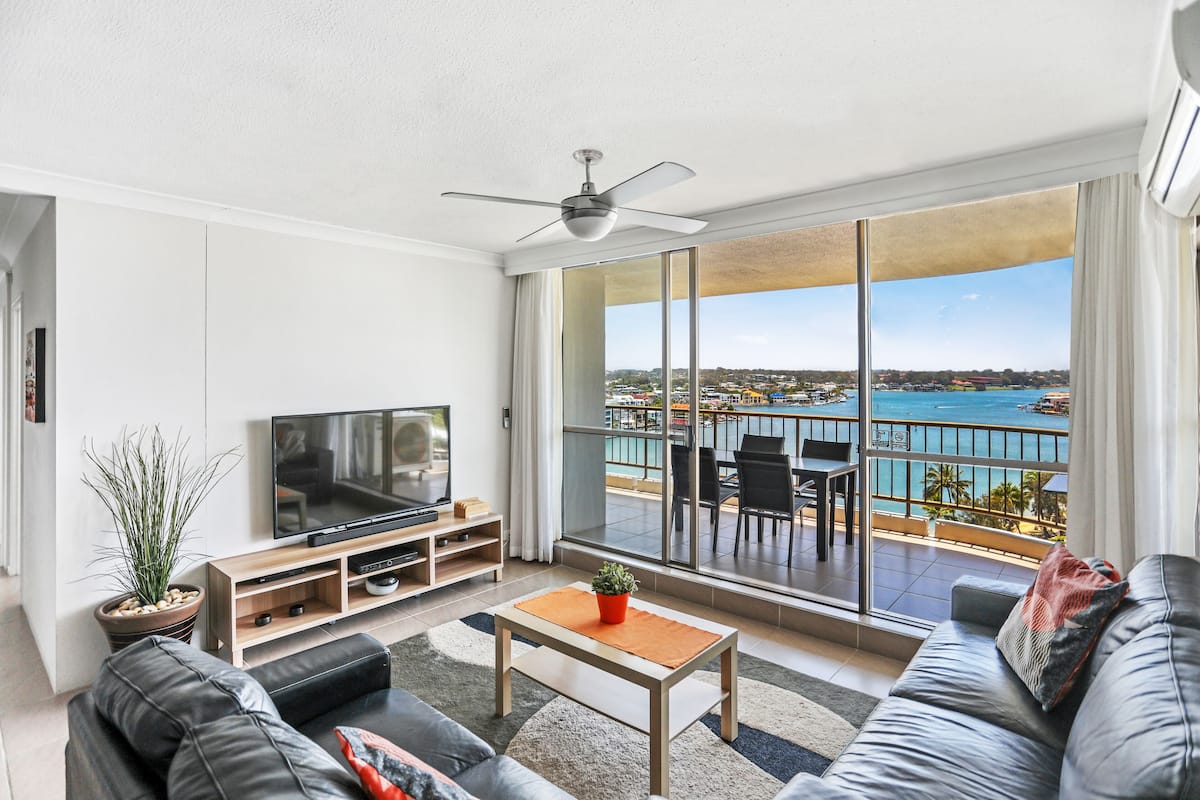 Una sala de estar moderna con un televisor grande, asientos cómodos, una mesita de centro y un balcón con vistas al mar.