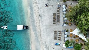 An aerial view of a beach with a boat docked on the shore, rows of sun loungers, umbrellas, and a small building with a sloped roof. - Trawangan Dive Centre (Gili Trawangan)