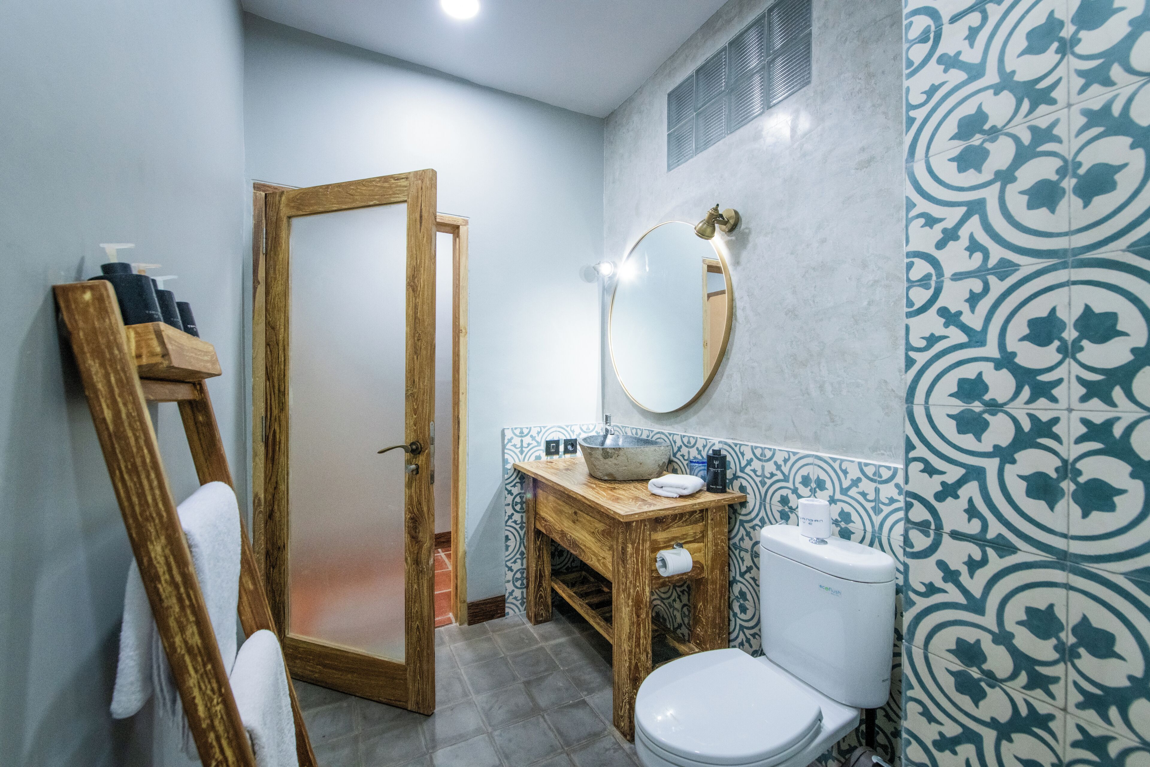 Un baño con tocador de madera, inodoro blanco, espejo y azulejos estampados.