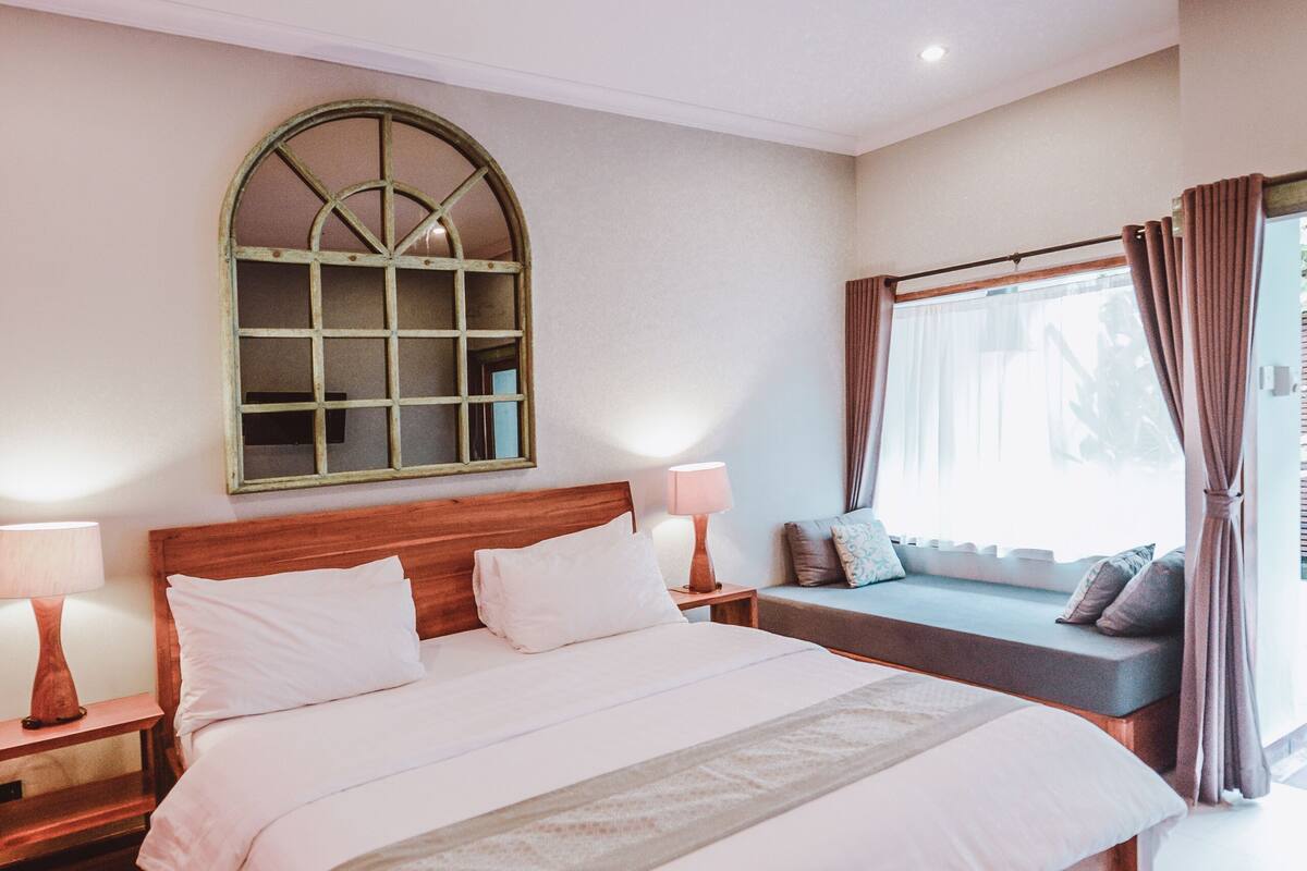 Ein Schlafzimmer mit einem großen Bett, einem Nachttisch mit Lampe, einem Fenster mit Vorhängen und einem an der Wand befestigten Spiegel.