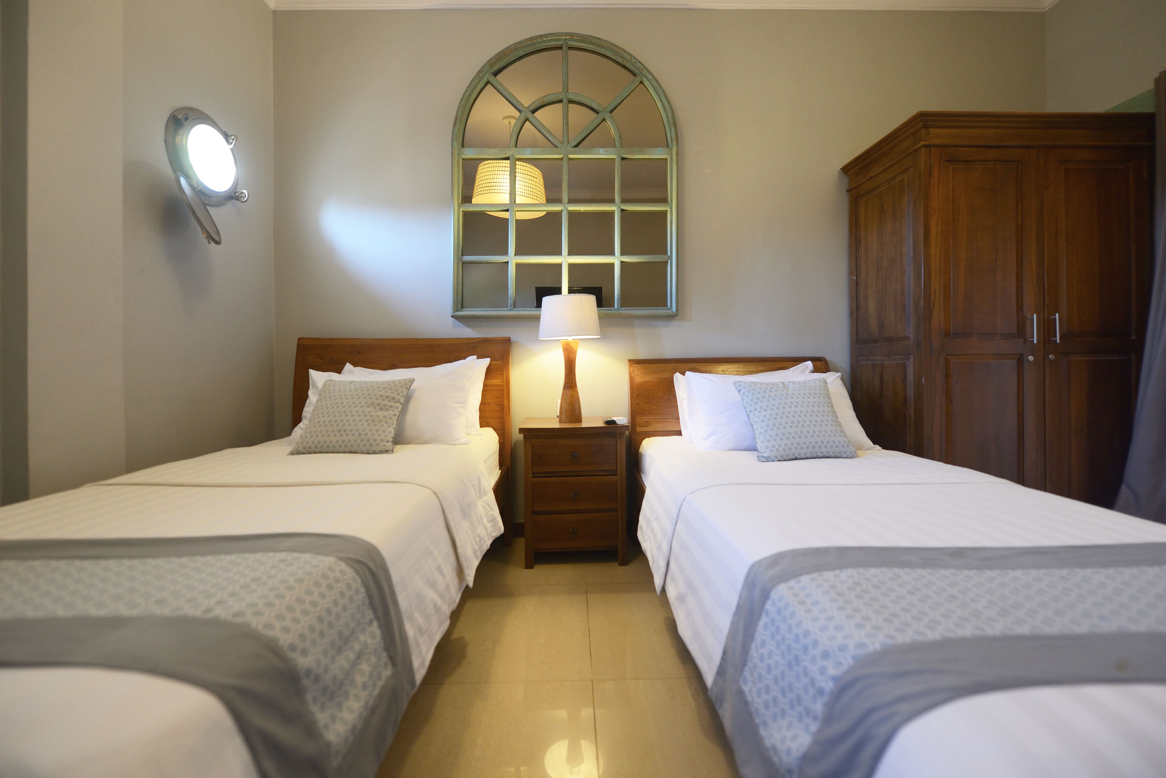 A hotel room with two beds, a nightstand with a lamp, a wooden wardrobe, and a window with a decorative frame.