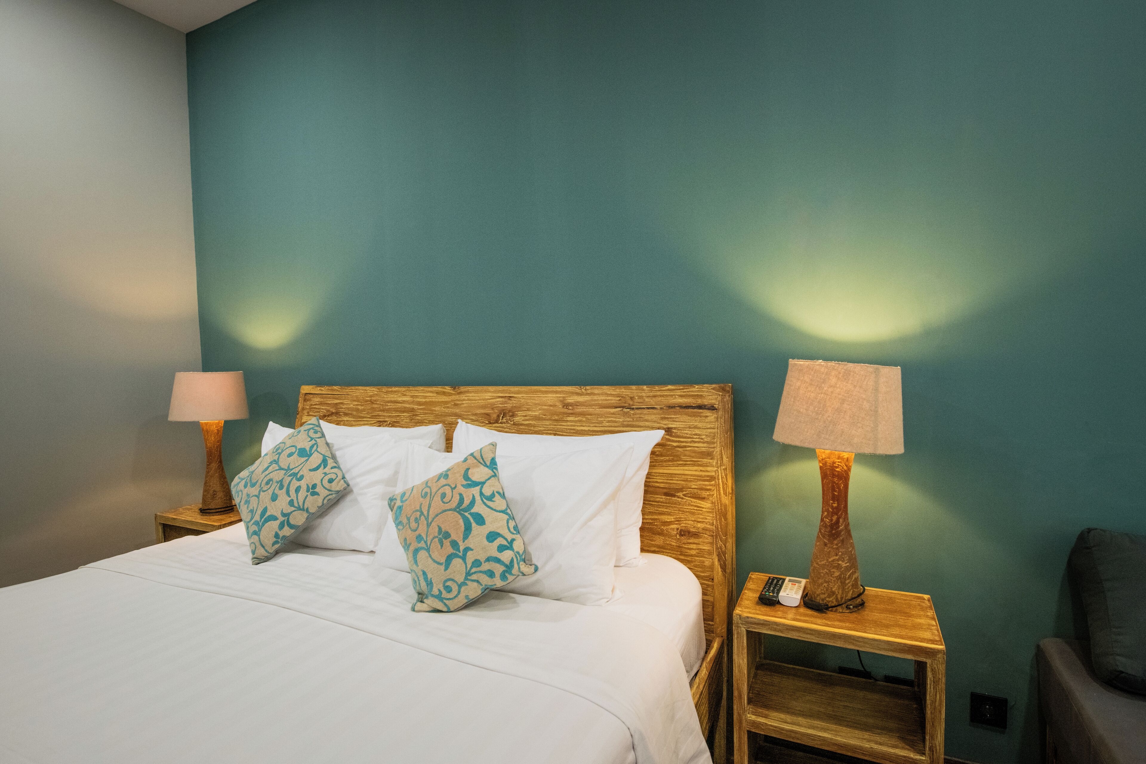 Un lit bien fait, avec du linge de lit blanc et deux coussins décoratifs, flanqué de deux tables de chevet en bois, sur lesquelles reposent des lampes et une télécommande.