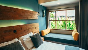 A bedroom with a large bed, a wooden headboard, a window seat with cushions, and a view of greenery outside. - Trawangan Dive Centre (Gili Trawangan)