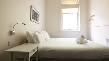 A neatly made bed with a white comforter and pillows, a bedside table with a lamp, a framed picture on the wall, and a window with a view of the sky.
