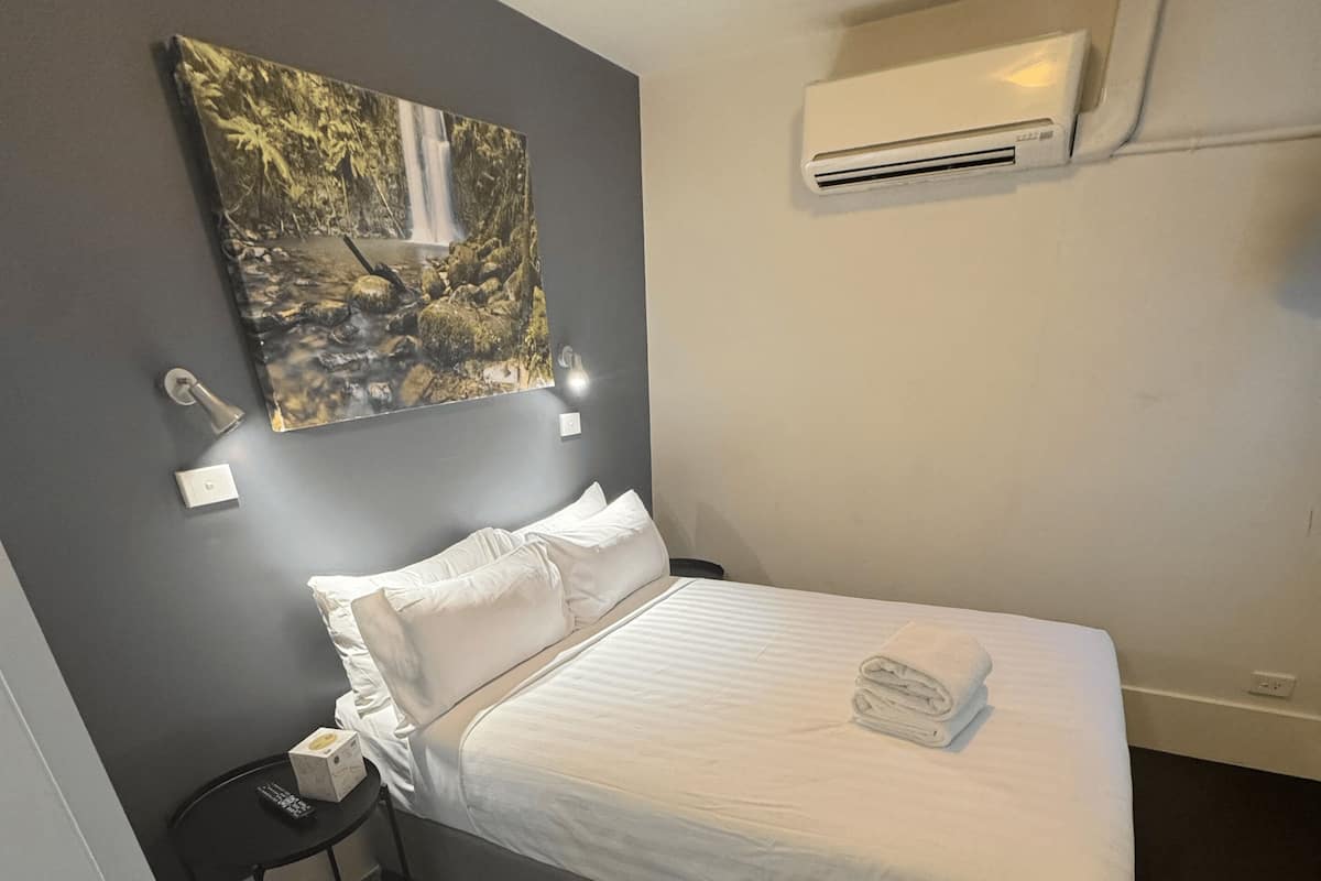 Cama bem arrumada com lençóis brancos, um controle remoto em uma mesinha ao lado e um ar condicionado fixado na parede, com um quadro acima da cama.