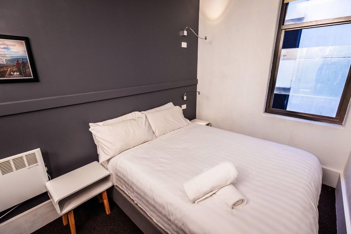 Uma cama bem arrumada com lençóis brancos, uma toalha enrolada, uma mesa de cabeceira, uma luminária na parede e uma janela com vista.