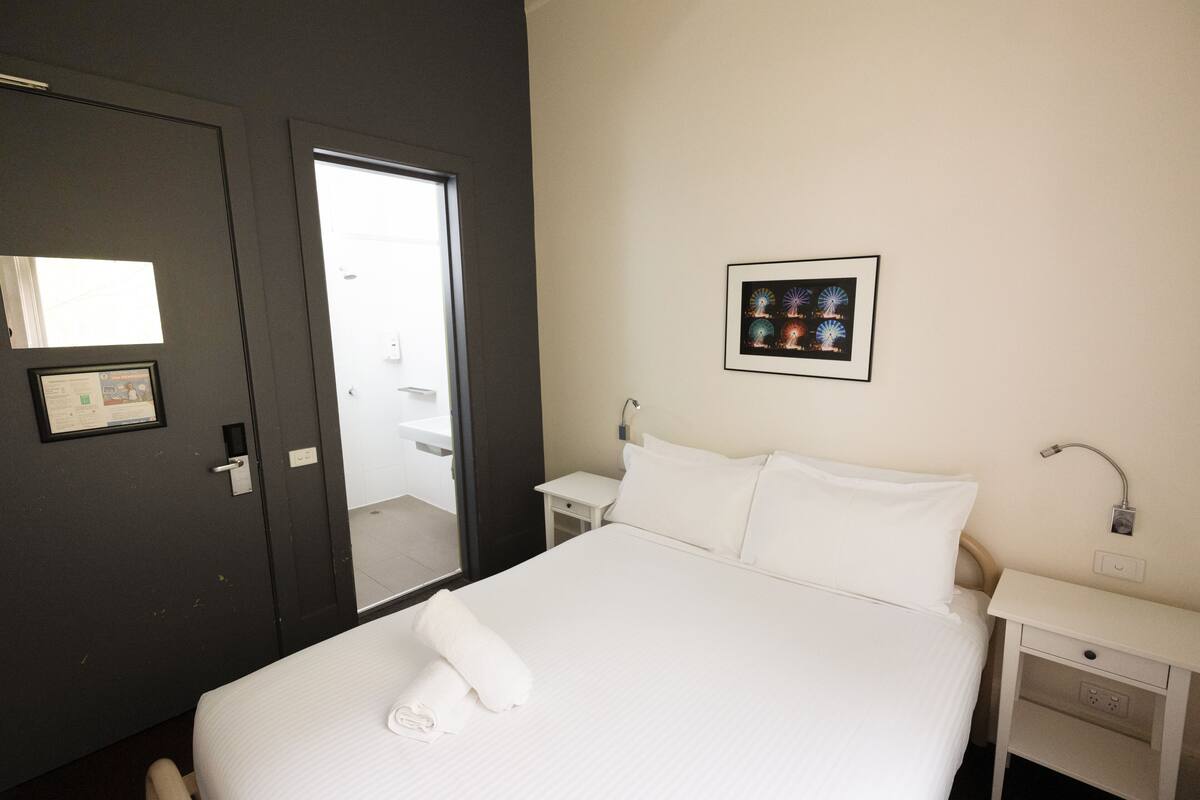 Quarto de hotel com cama, mesa de cabeceira, quadro na parede e porta para o banheiro.
