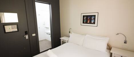 Una habitación de hotel con una cama, una mesita de noche, un cuadro en la pared y una puerta que da a un baño.