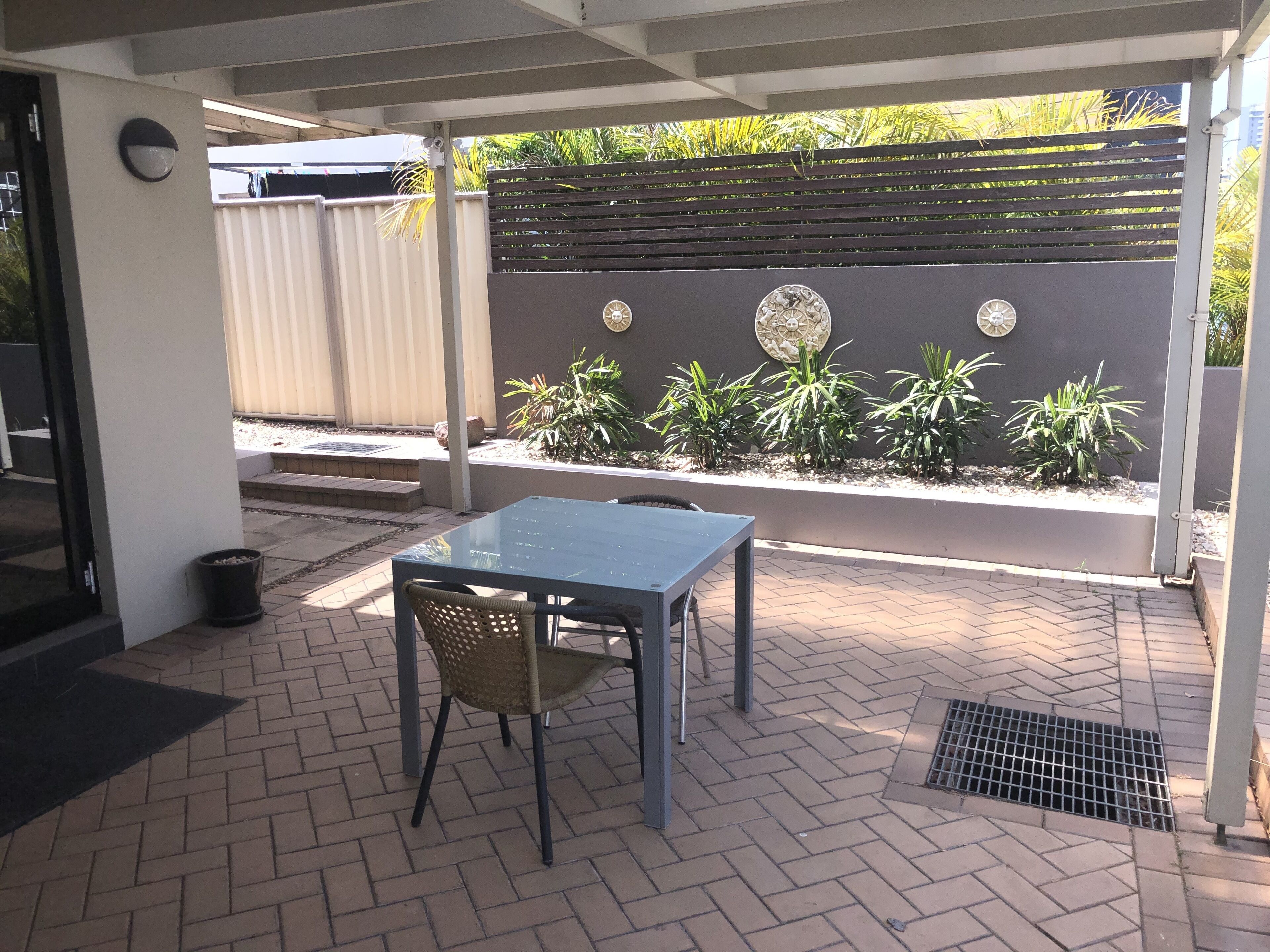 Un espace extérieur aménagé en patio, avec une table et des chaises, une poubelle en métal et un mur décoratif agrémenté de plantes et d’œuvres d’art mural.