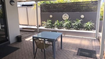 Un área de patio al aire libre con una mesa y una silla, un bote de basura metálico y una pared decorativa con plantas y adornos.