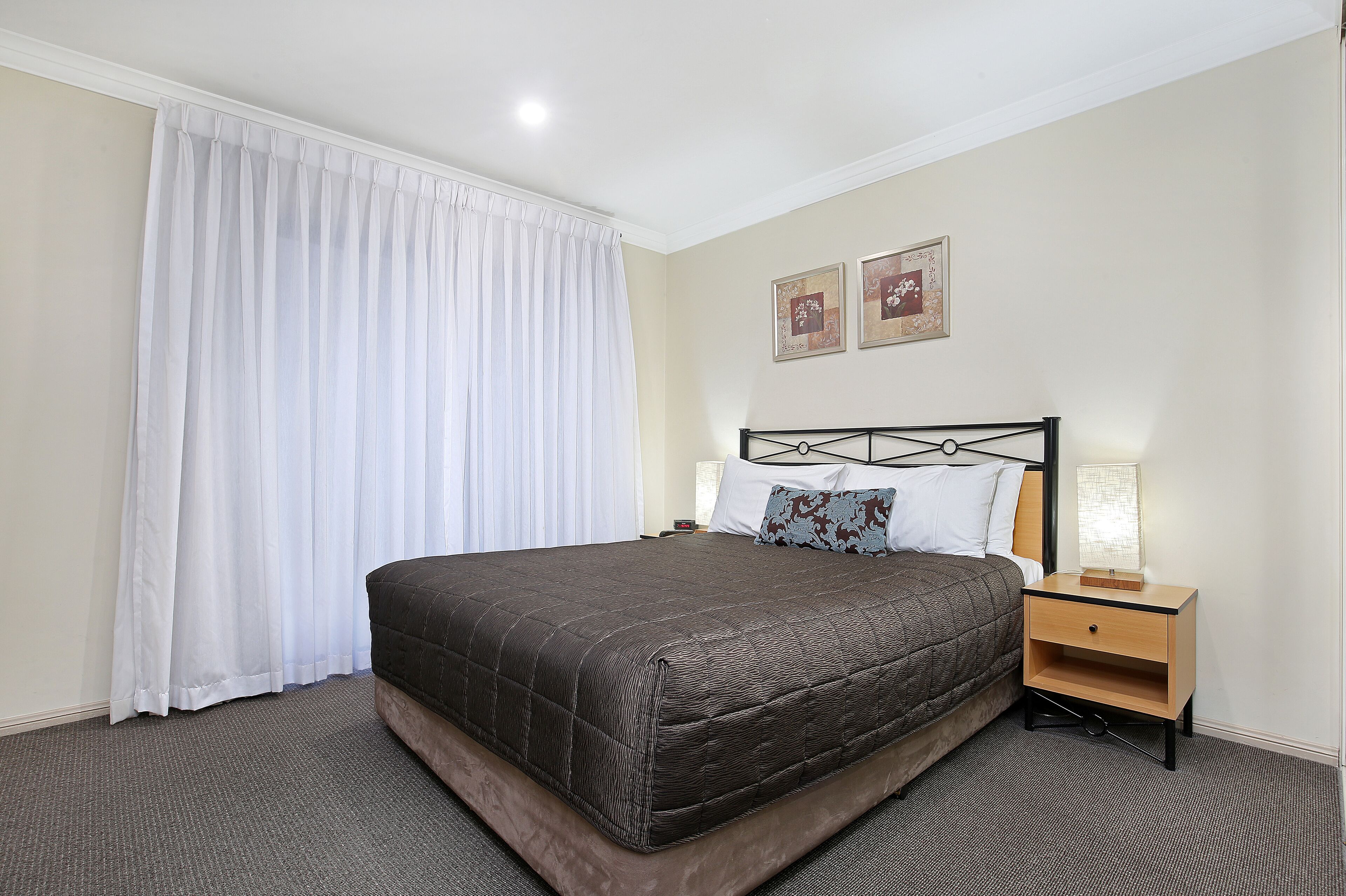A neatly made bed with a dark quilt, a bedside table with a lamp, and a large window with white curtains.