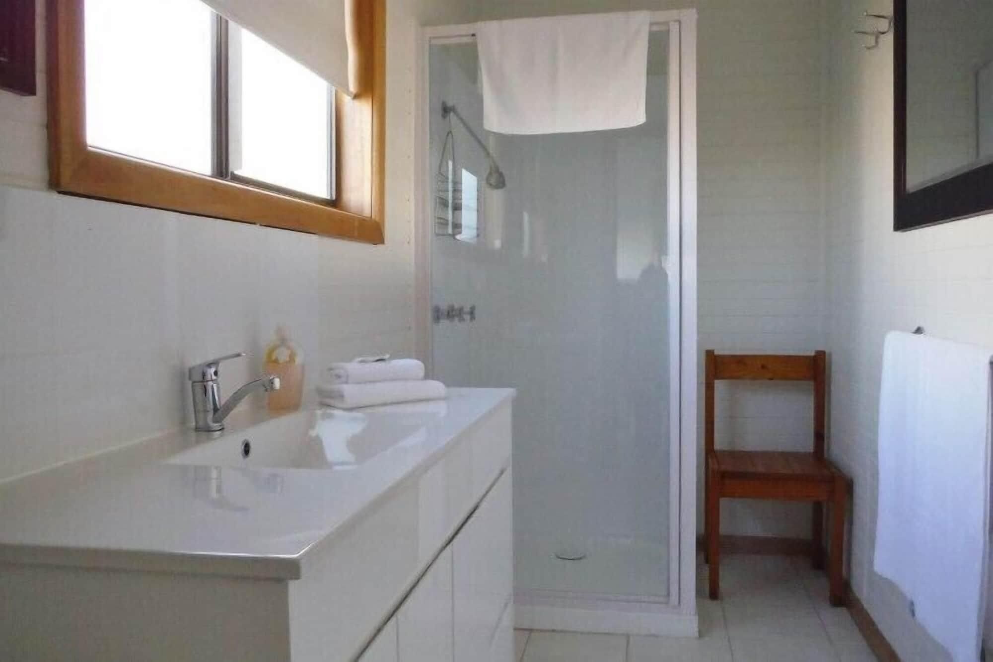 Ein Badezimmer mit einem weißen Waschbecken, einem Spiegel, einer Dusche und einem Holzstuhl.
