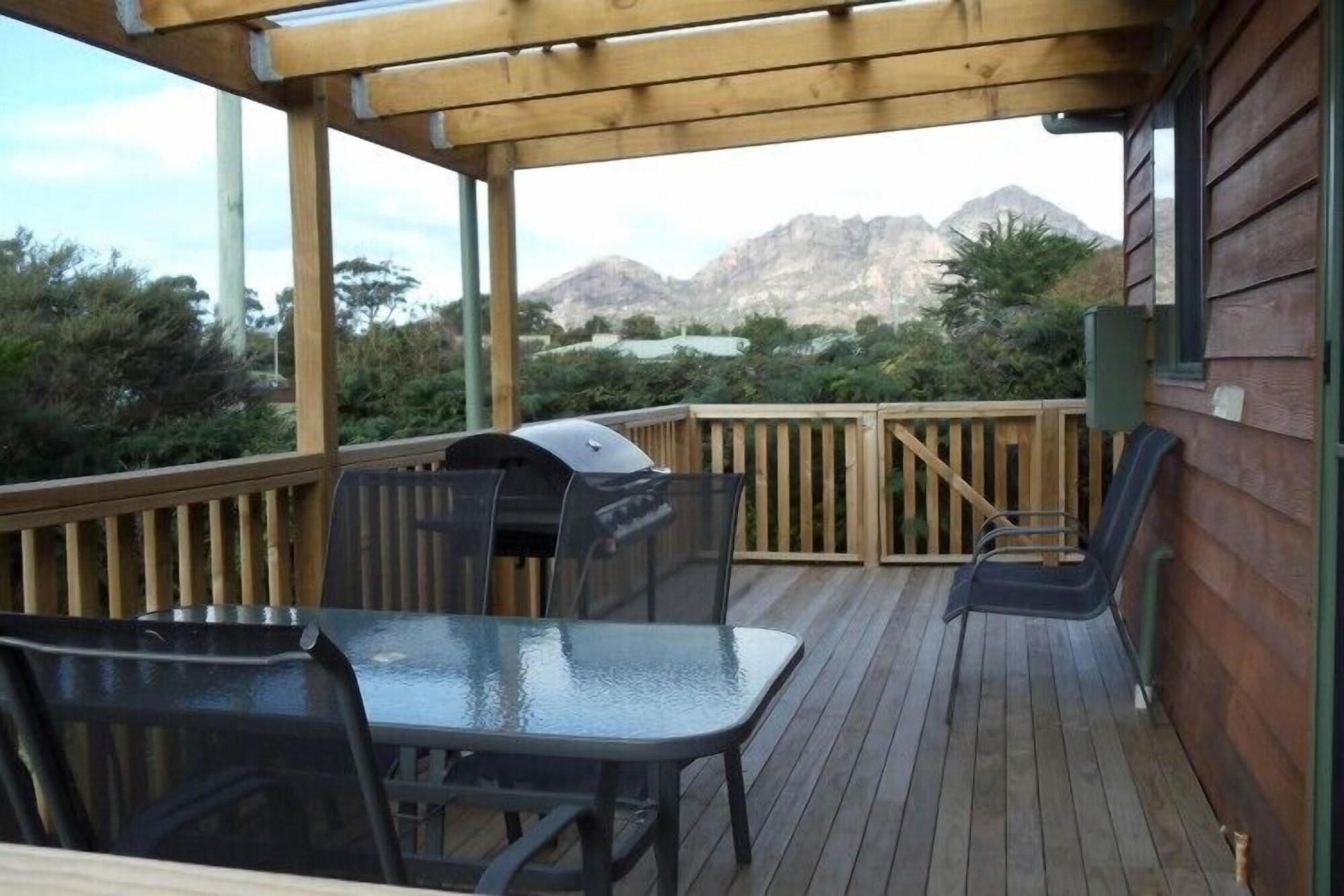 Una terraza de madera con una parrilla, mesa y sillas, con vista a un paisaje montañoso.