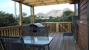 Una terraza de madera con una parrilla, mesa y sillas, con vista a un paisaje montañoso.