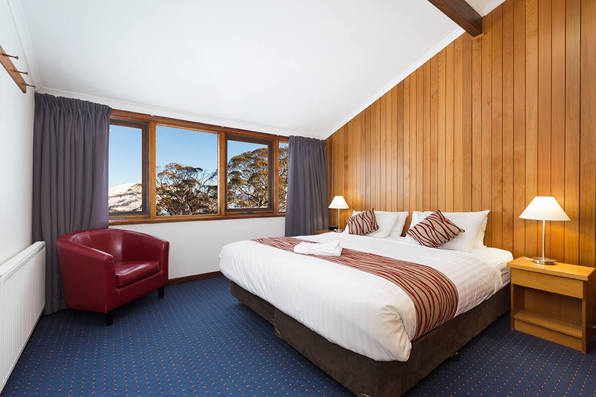 Habitación de hotel con una cama grande, una silla roja, dos mesitas de noche con lámparas y vistas a árboles a través de amplias ventanas.