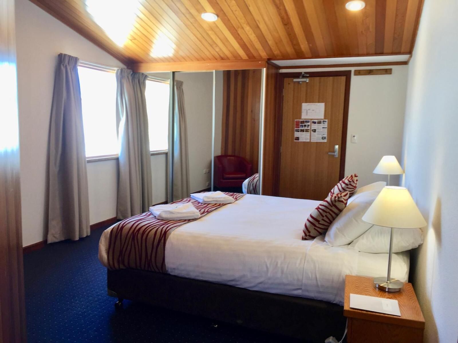 A hotel room with a bed, a nightstand, a lamp, a chair, and a door with a notice.