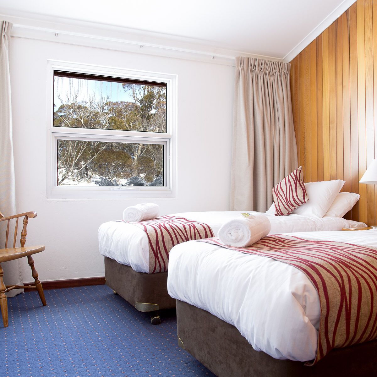 A hotel room with two beds, a wooden chair, a window with a view of trees, and a curtain.