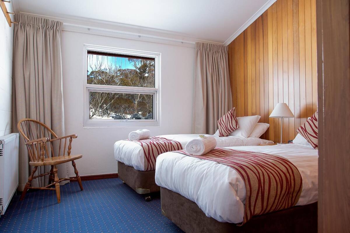 Habitación de hotel con dos camas, cabecero de madera, ventana con cortinas, una silla y un radiador.