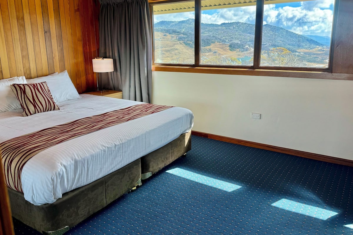 Un dormitorio con una cama grande, una mesita de noche, una lámpara y vistas a montañas a través de una ventana.