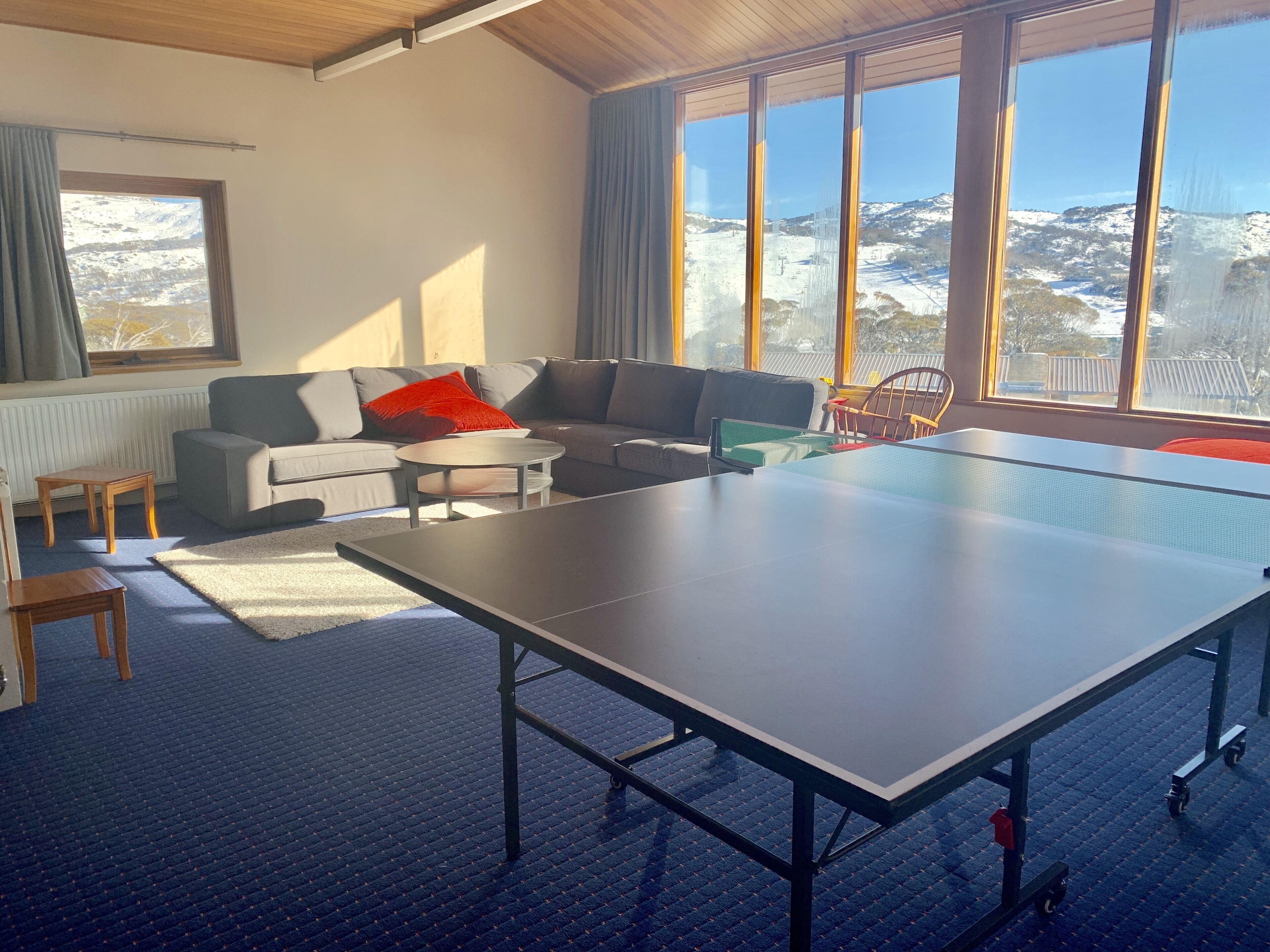 A living room with a ping pong table, grey sofas, and a view of a snowy landscape through large windows.