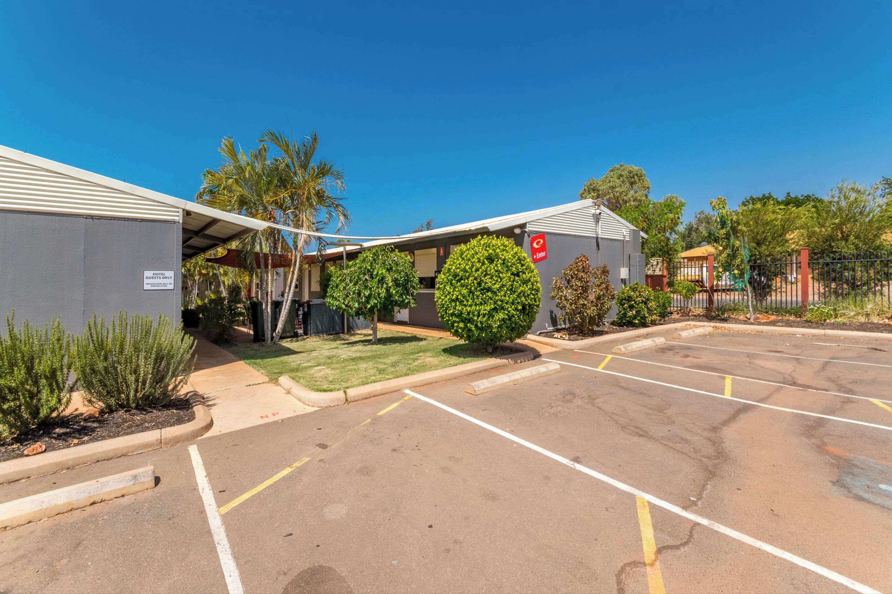 A hotel with a covered parking area, a sign indicating a motel, and a fenced garden area with trees and shrubs.