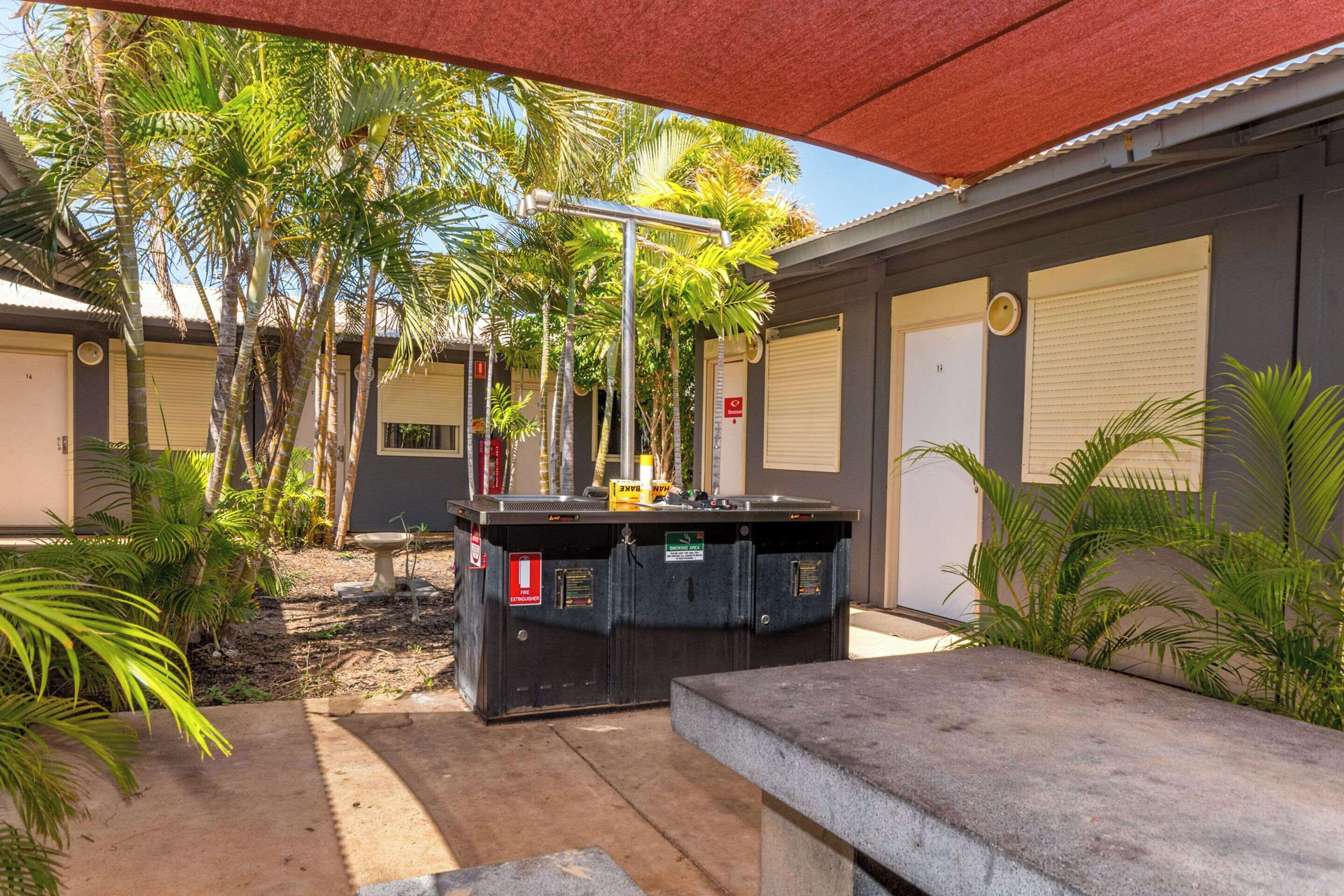 A covered outdoor area with a pool table, palm trees, and a building with closed shutters.