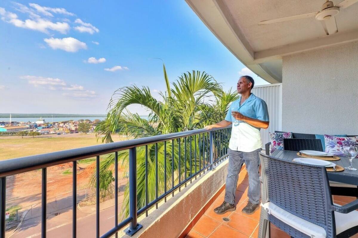 A balcony with a view of a road and palm trees, a table with chairs, and a ceiling fan.