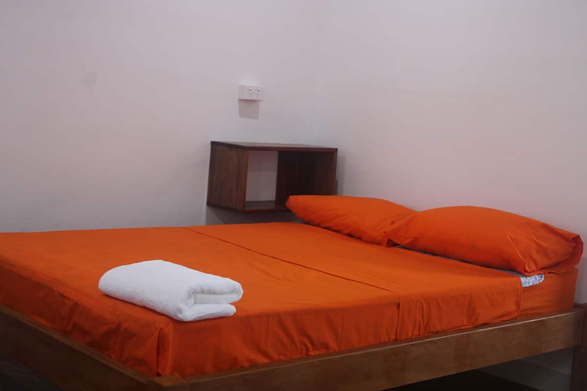 Een eenpersoonsbed met een oranje sprei en kussens, een houten hoofdbord en een klein nachtkastje met een stopcontact.