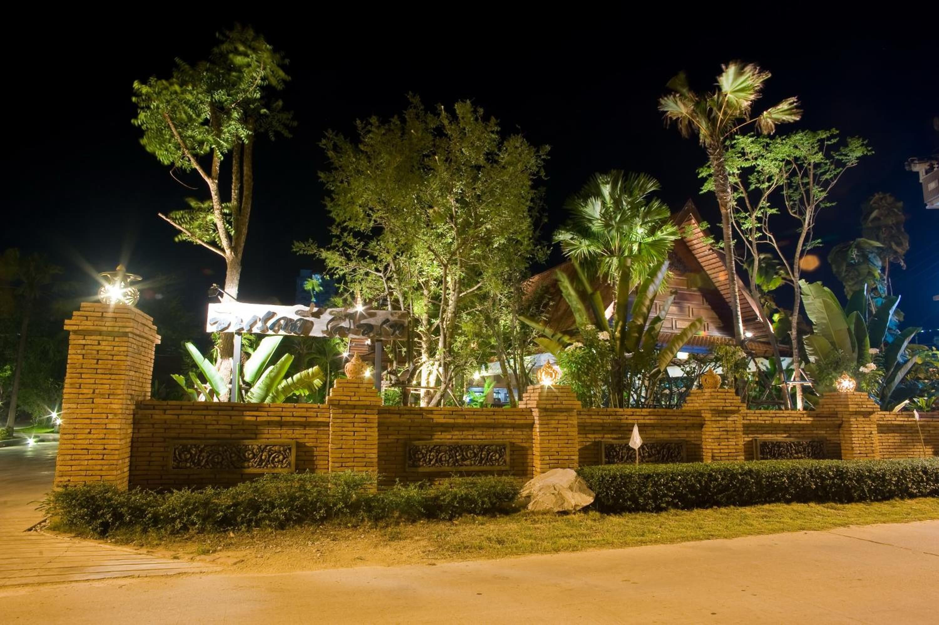 Una entrada bien iluminada con pared de ladrillo, detalles metálicos decorativos y abundante vegetación, incluyendo palmeras.