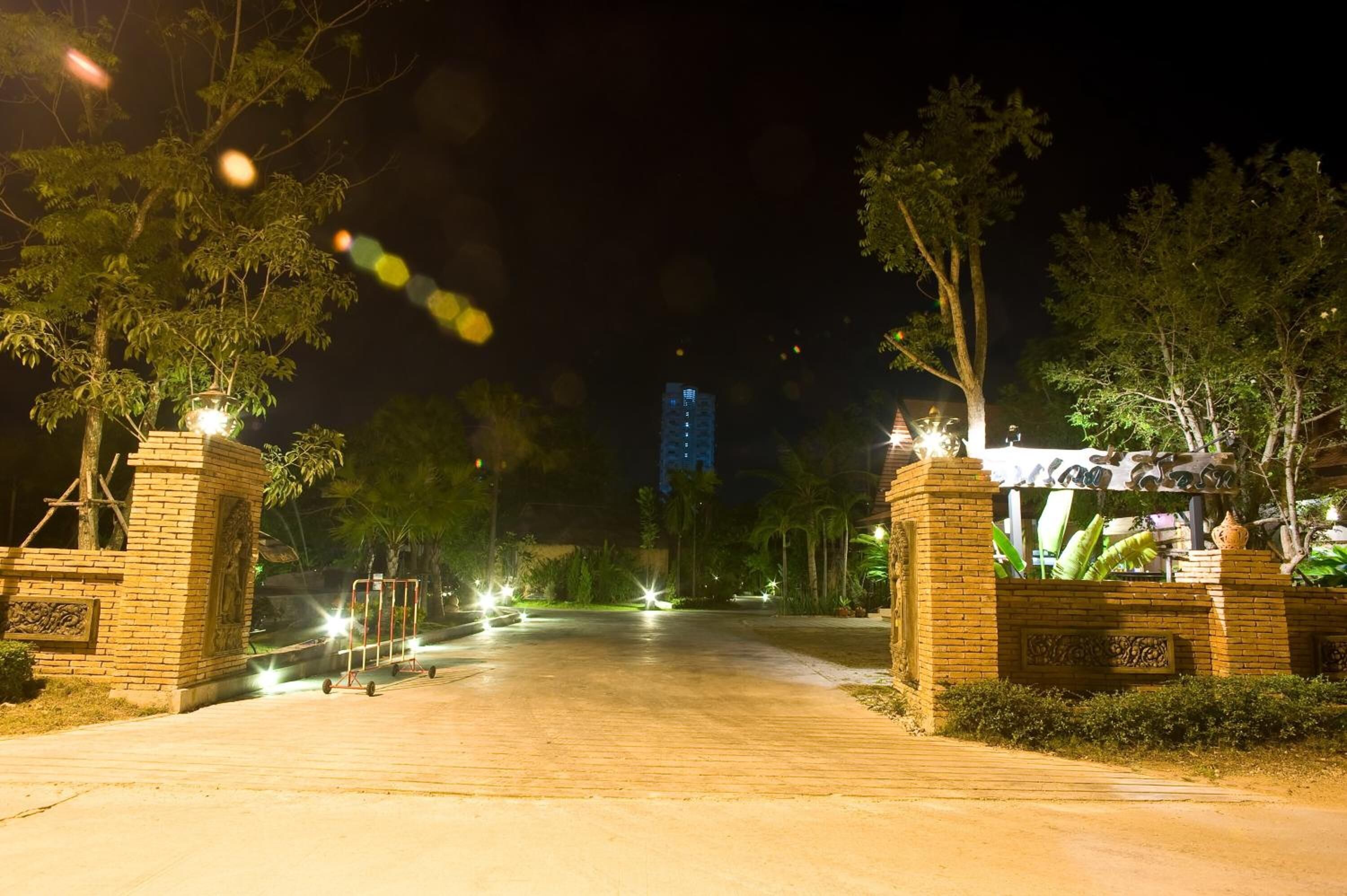 Une vue nocturne d’une entrée fermée par un portail, avec des allées illuminées, entourée d’arbres et d’un bâtiment situé au loin.
